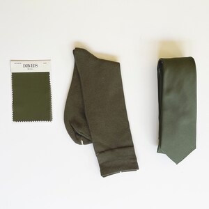 May include: An overhead shot of a coordinated set of accessories in a muted olive green color. The set includes a necktie, a pair of socks, and a color swatch from David's Bridal. The items are arranged on a white surface.