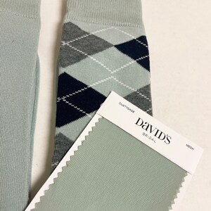 May include: A pair of light sage green socks featuring a dark navy blue and grey argyle pattern with white stitching details. Next to the socks is a rectangular fabric swatch labelled DUSTYSAGE DAVID'S BRIDAL MESH in a light sage green colour.