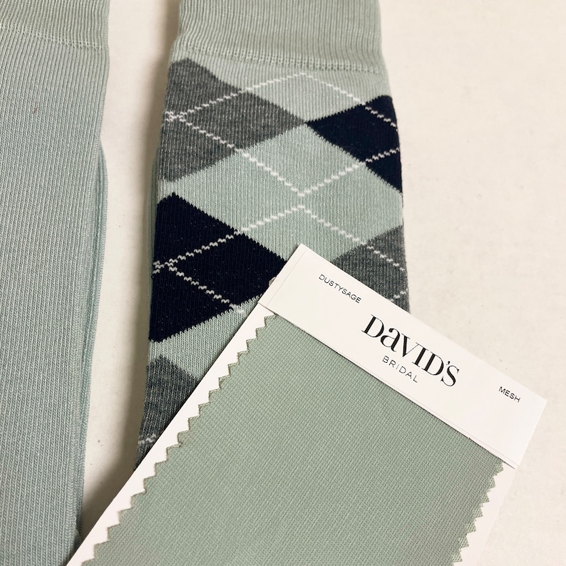 May include: A pair of light sage green socks featuring a dark navy blue and grey argyle pattern with white stitching details. Next to the socks is a rectangular fabric swatch labelled DUSTYSAGE DAVID'S BRIDAL MESH in a light sage green colour.
