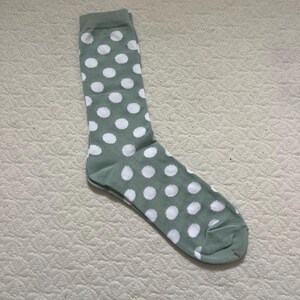 May include: A sage green crew-length sock featuring a repeating pattern of white polka dots. The sock has a ribbed cuff at the top, a defined heel section, and a rounded toe area, all constructed from a soft, knit fabric with a consistent colour.