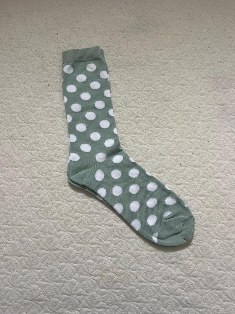 May include: A sage green crew-length sock featuring a repeating pattern of white polka dots. The sock has a ribbed cuff at the top, a defined heel section, and a rounded toe area, all constructed from a soft, knit fabric with a consistent colour.