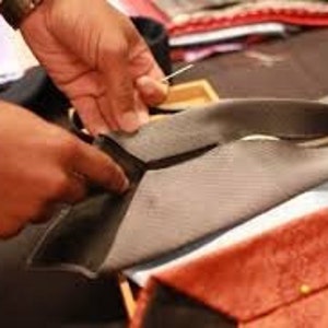 May include: A person's hands are shown working on a gray tie, with a needle and thread visible. The tie is being sewn, with other ties in various colors and patterns in the background. The ties appear to be made of silk or a similar material.