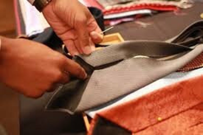 May include: A person's hands are shown working on a gray tie, with a needle and thread visible. The tie is being sewn, with other ties in various colors and patterns in the background. The ties appear to be made of silk or a similar material.