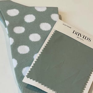 May include: A sage green sock with a white polka dot pattern and a rectangular fabric swatch card labelled David's Bridal in the colour Dusty Sage. The swatch is made of a solid sage green mesh fabric with pinked edges and a white paper header.