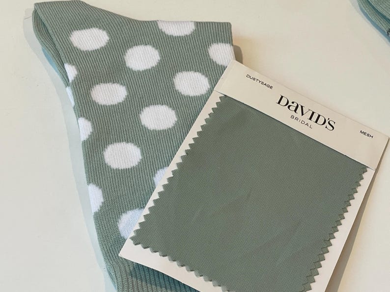 May include: A sage green sock with a white polka dot pattern and a rectangular fabric swatch card labelled David's Bridal in the colour Dusty Sage. The swatch is made of a solid sage green mesh fabric with pinked edges and a white paper header.
