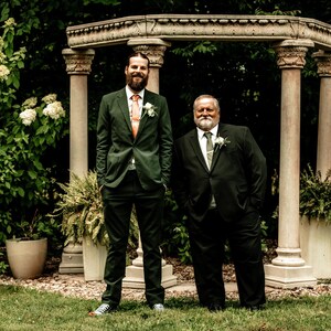 May include: Two men in suits pose outdoors. One wears a dark green suit with an orange tie, the other a black suit with a green tie. They stand in front of a stone structure with columns, surrounded by greenery and potted plants.
