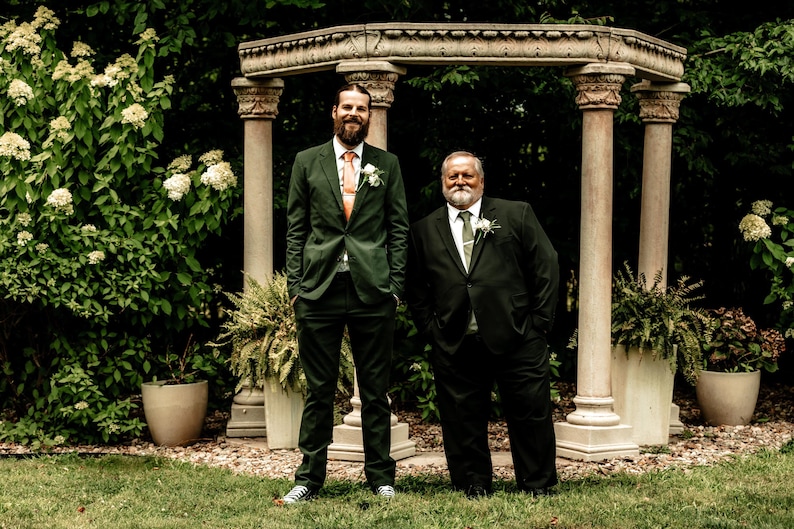 May include: Two men in suits pose outdoors. One wears a dark green suit with an orange tie, the other a black suit with a green tie. They stand in front of a stone structure with columns, surrounded by greenery and potted plants.