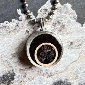 May include: A silver chain necklace with a round pendant. The pendant is made of metal and has a black and brown resin inlay with a circular design.