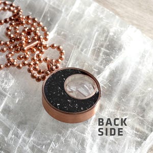 May include: A copper chain necklace with a black and white resin pendant. The pendant features a crescent moon shape with a white crystal inside. The text "BACK SIDE" is visible on the surface below the pendant.