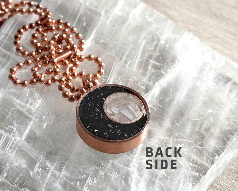 May include: A copper chain necklace with a black and white resin pendant. The pendant features a crescent moon shape with a white crystal inside. The text "BACK SIDE" is visible on the surface below the pendant.
