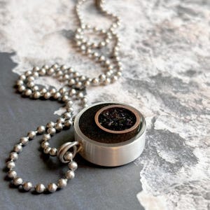 May include: A silver chain necklace with a round pendant. The pendant is made of metal and has a black resin inlay with a copper circle in the center. The copper circle is filled with black specks.