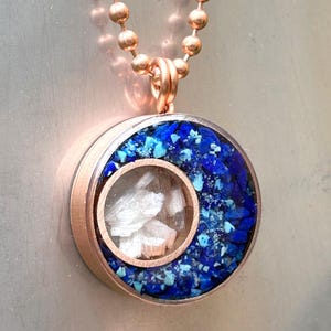 May include: A rose gold necklace with a round pendant featuring a clear window revealing white crystals and a blue and turquoise resin background.