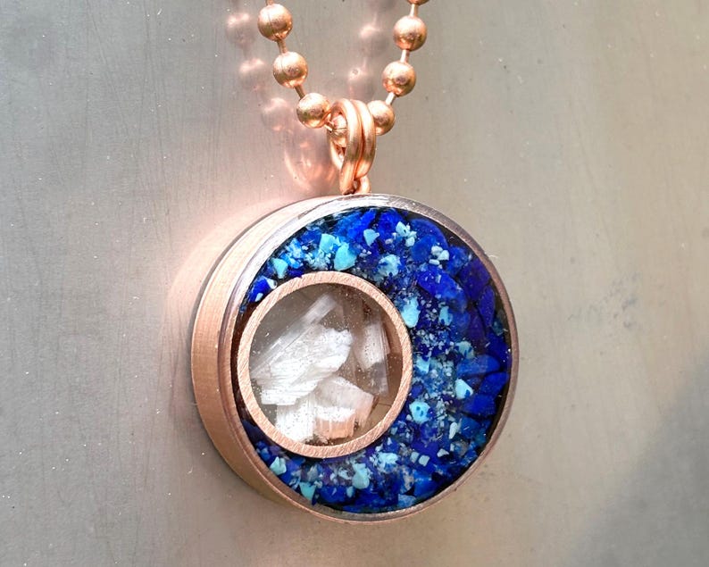 May include: A rose gold necklace with a round pendant featuring a clear window revealing white crystals and a blue and turquoise resin background.