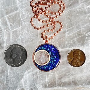 May include: A copper chain necklace with a pendant featuring a blue and white crescent moon design. The pendant is made of resin and has a clear center with a white stone. The necklace is on a white background.