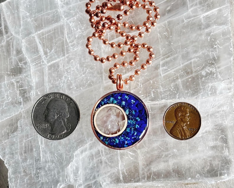 May include: A copper chain necklace with a pendant featuring a blue and white crescent moon design. The pendant is made of resin and has a clear center with a white stone. The necklace is on a white background.
