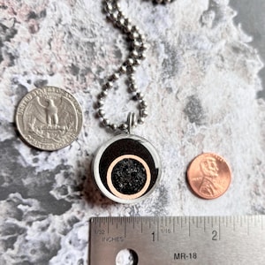May include: A silver chain necklace with a round pendant. The pendant is made of metal and has a black background with a copper ring and a black and silver speckled center. The pendant is about the size of a US quarter.