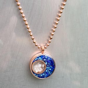 May include: A rose gold necklace with a round pendant featuring a blue and white resin design. The pendant has a crescent moon shape with white resin inside.
