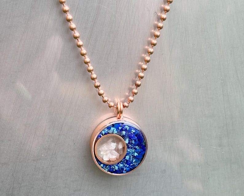 May include: A rose gold necklace with a round pendant featuring a blue and white resin design. The pendant has a crescent moon shape with white resin inside.