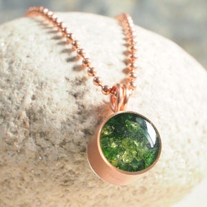May include: A rose gold pendant necklace featuring a round, bezel-set green stone. The stone has a textured appearance. The necklace chain is a matching rose gold ball chain. The pendant is displayed on a light-colored, textured stone.
