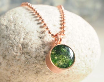 May include: A rose gold pendant necklace featuring a round, bezel-set green stone. The stone has a textured appearance. The necklace chain is a matching rose gold ball chain. The pendant is displayed on a light-colored, textured stone.