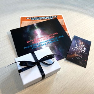 May include: A white gift box with a black ribbon is on a wooden surface. The box is surrounded by three cards with text that reads "We are living in a sea of electrosmog", "With stardust activated we rise together, fierce and unwavering in love, virtue and strength." and "Stardust Alchemy".