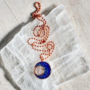 May include: A copper chain necklace with a crescent moon pendant. The pendant is made of copper and has a blue and white crystal inside. The necklace is displayed on a white selenite slab.
