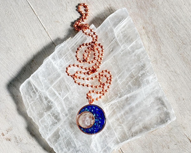 May include: A copper chain necklace with a crescent moon pendant. The pendant is made of copper and has a blue and white crystal inside. The necklace is displayed on a white selenite slab.
