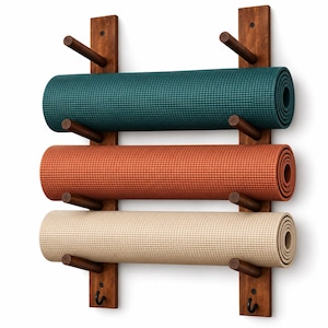 May include: A wall-mounted wooden rack with two vertical supports and six horizontal pegs holding three rolled yoga mats in teal, terracotta, and beige. The rack features two small black metal hooks at the bottom for additional storage.