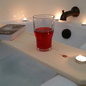 K&ouml;nnte beinhalten: Ein Holz-Badetablett mit einem Buch, einem Glas roten Fl&uuml;ssigkeit und mehreren brennenden Kerzen. Das Tablett liegt am Rand einer wei&szlig;en Badewanne.