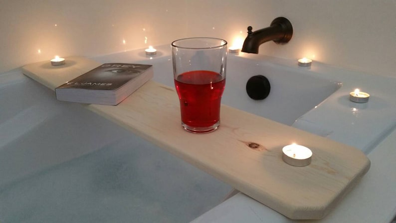 K&ouml;nnte beinhalten: Ein Holz-Badetablett mit einem Buch, einem Glas roten Fl&uuml;ssigkeit und mehreren brennenden Kerzen. Das Tablett liegt am Rand einer wei&szlig;en Badewanne.