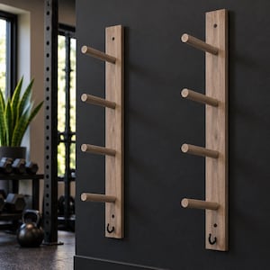 May include: Two vertical light brown wooden wall racks, each featuring four horizontal cylindrical pegs and a small black metal hook at the bottom, mounted on a dark wall for storage of gym equipment or accessories.