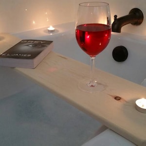K&ouml;nnte beinhalten: Ein Holz-Badetablett mit einem Glas Rotwein, einem Buch mit dem Titel "GREY" von E.L. James und mehreren brennenden Kerzen. Das Tablett liegt am Rand einer wei&szlig;en Badewanne.