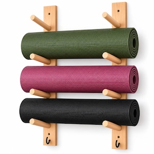 May include: A wall-mounted light wood storage rack featuring two vertical panels with six horizontal pegs holding three rolled yoga mats in olive green, magenta, and black. Two small black metal hooks are attached to the bottom of the rack.