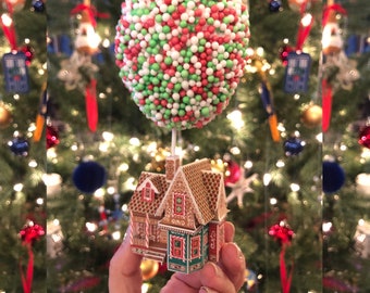May include: A festive Christmas tree ornament featuring a miniature gingerbread house with red, green, and gold accents. Above it, a lollipop-shaped treat covered in red, green, and white sprinkles. The background is a blurred Christmas tree with twinkling lights.