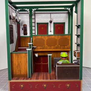 May include: A miniature papercraft model of a building interior with a green trim, wooden walls, and a red brick floor. A green frog figurine sits at a desk with a cup of coffee. The building has a red door, a telephone, and a sign that reads "Doors Open 9-5".