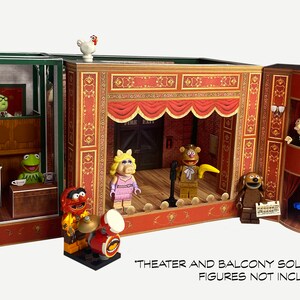 May include: A three-section cardboard playset depicting a Muppet theater stage with a red curtain, a balcony, and a backstage area. The set is decorated with gold trim and features a variety of Muppet characters, including Miss Piggy, Fozzie Bear, Animal, and Gonzo. The text "THEATER AND BALCONY SOLD SEPARATELY FIGURES NOT INCLUDED" is printed at the bottom of the image.