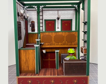 May include: A miniature dollhouse with a green frame, brown wood paneling, and a red brick base. The interior features a wooden counter, a telephone, and a green frog figurine. The dollhouse is decorated with a red and white checkered floor and a red brick base.