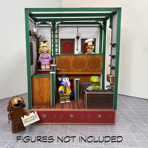 May include: A green and brown papercraft model of a Muppet Show set. The set features a stage, a counter, and a back wall with a brick pattern. The model is empty, but it includes a sign that says "Figures not included".