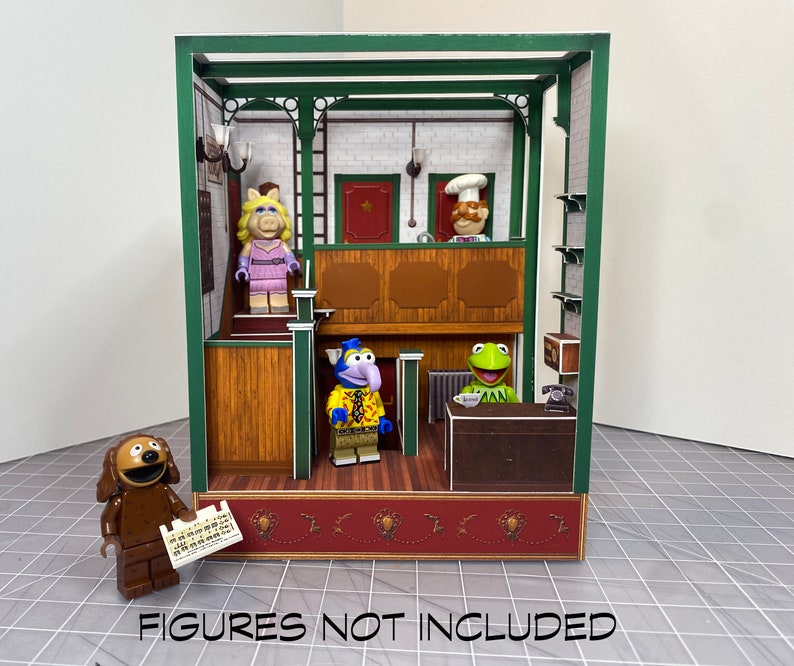 May include: A green and brown papercraft model of a Muppet Show set. The set features a stage, a counter, and a back wall with a brick pattern. The model is empty, but it includes a sign that says "Figures not included".