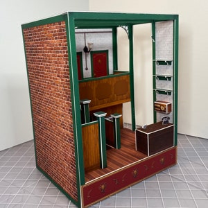 May include: A miniature paper model of a building interior with a brick exterior, green trim, and a wooden interior. The interior features a balcony, a counter, and shelves.