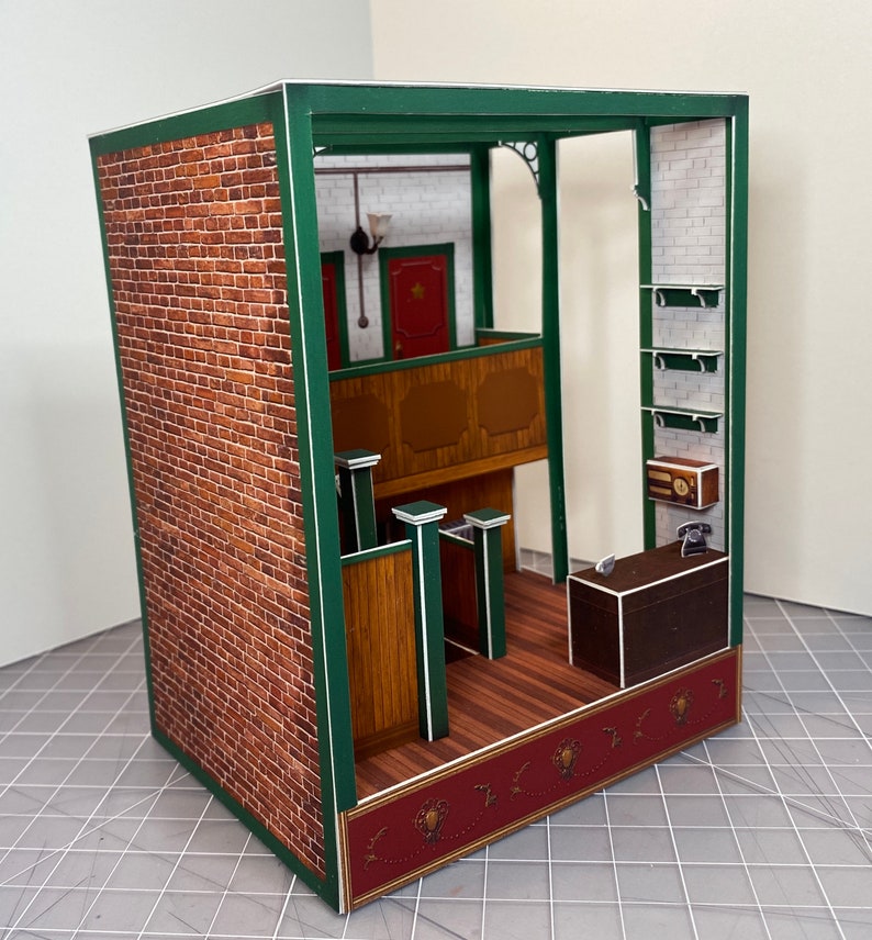 May include: A miniature paper model of a building interior with a brick exterior, green trim, and a wooden interior. The interior features a balcony, a counter, and shelves.