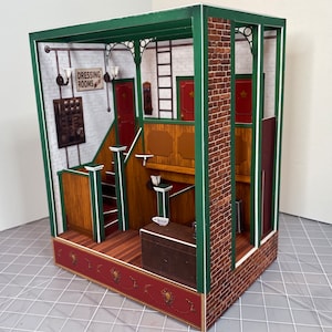 May include: A miniature paper model of a building interior with a green frame, brick wall, and wooden stairs. The sign above the stairs reads "Dressing Rooms".
