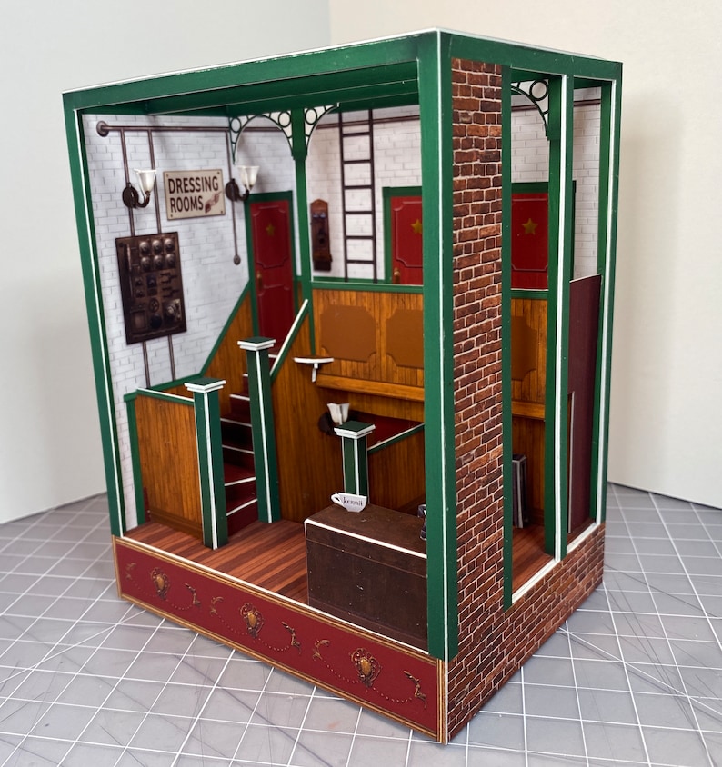 May include: A miniature paper model of a building interior with a green frame, brick wall, and wooden stairs. The sign above the stairs reads "Dressing Rooms".