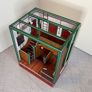 May include: A paper model of a building with a green frame and white trim. The building has a staircase, a counter, and a sign that says "Dress Rooms".
