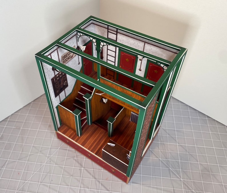 May include: A paper model of a building with a green frame and white trim. The building has a staircase, a counter, and a sign that says "Dress Rooms".