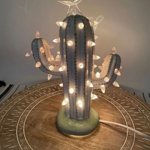 May include: A ceramic cactus Christmas tree with a clear star topper and string lights. The cactus is light blue with green accents and a green base. The lights are small and clear, and the tree is plugged into a white cord.