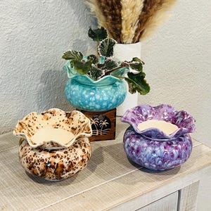 May include: Three ceramic wax warmers in a row. The first is brown with a speckled pattern, the second is turquoise, and the third is purple. Each warmer has a ruffled top and a round base. A vase of dried pampas grass is in the background.