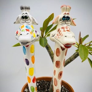 May include: Two ceramic giraffe figurines with painted spots, one with rainbow spots and the other with brown spots, are in a terracotta pot with a green plant.