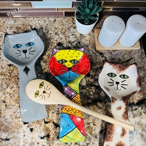 May include: Set of three ceramic cat-shaped spoon rests. One is gray with black eyes, one is multi-colored with a patchwork design, and one is white with brown spots.