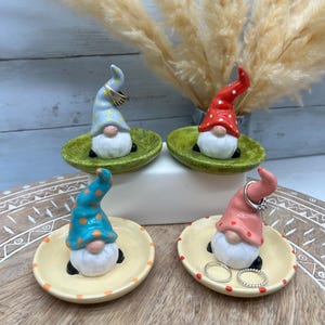May include: Four ceramic gnome ring holders. Each gnome has a different colored hat with polka dots. The ring holders are in the shape of small dishes with a decorative border. Rings are displayed on two of the holders.